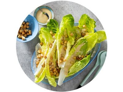 Romaine lettuce leaves topped with croutons and dressing are arranged on a blue plate, with extra croutons, dressing, and utensils placed nearby on a gray surface.