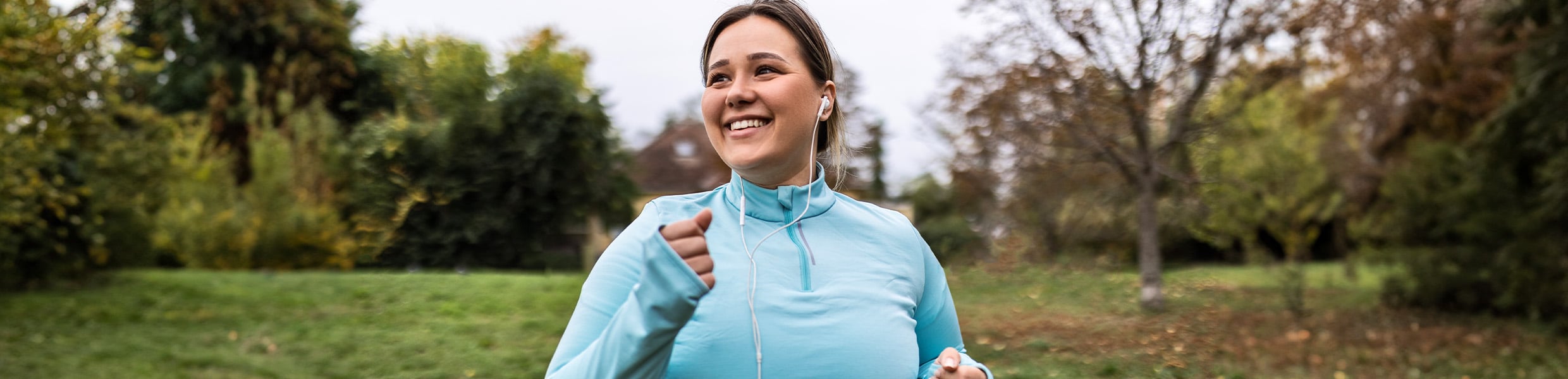Person wearing a light blue athletic top and earphones walks through a park with trees and grass in the background.