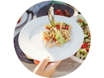 Pasta salad with rotini, lettuce, and diced vegetables is being served onto a white plate from a larger salad bowl in an outdoor dining setting.