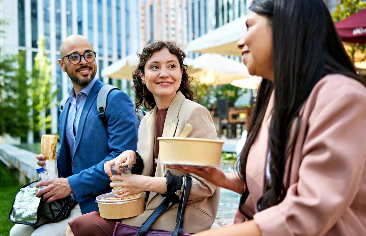 “Three adults sitting outdoors near office buildings, eating takeout meals from containers and holding drinks while conversing at a city plaza.”